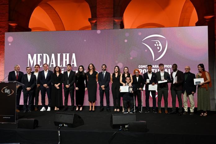 Seventeen people wearing black suits or dresses pose for a photograph in front of a purple banner with a logo of the National Paralympic Committee of Portugal