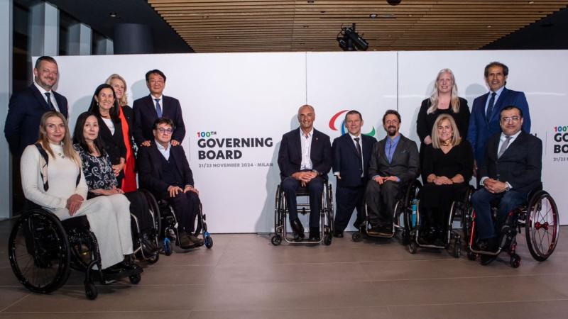 Fourteen people wearing formal clothing are posing in front of a banner that says "100th Governing Board"