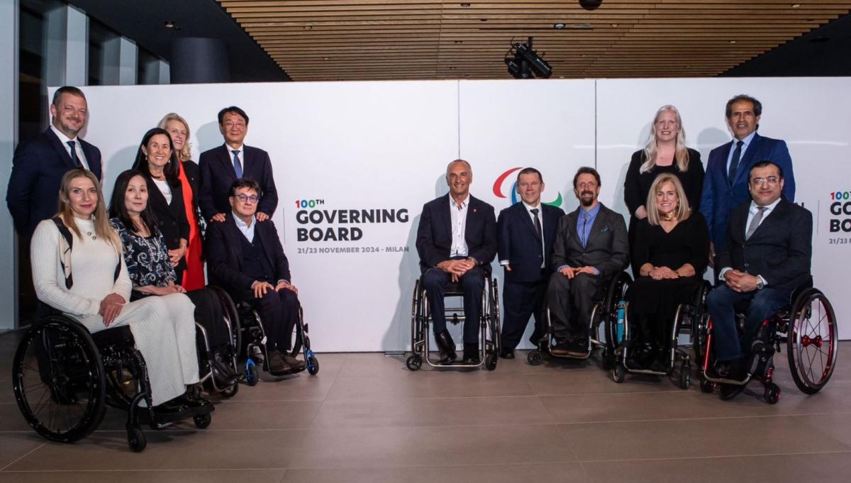 Fourteen people wearing formal clothing are posing in front of a banner that says "100th Governing Board"