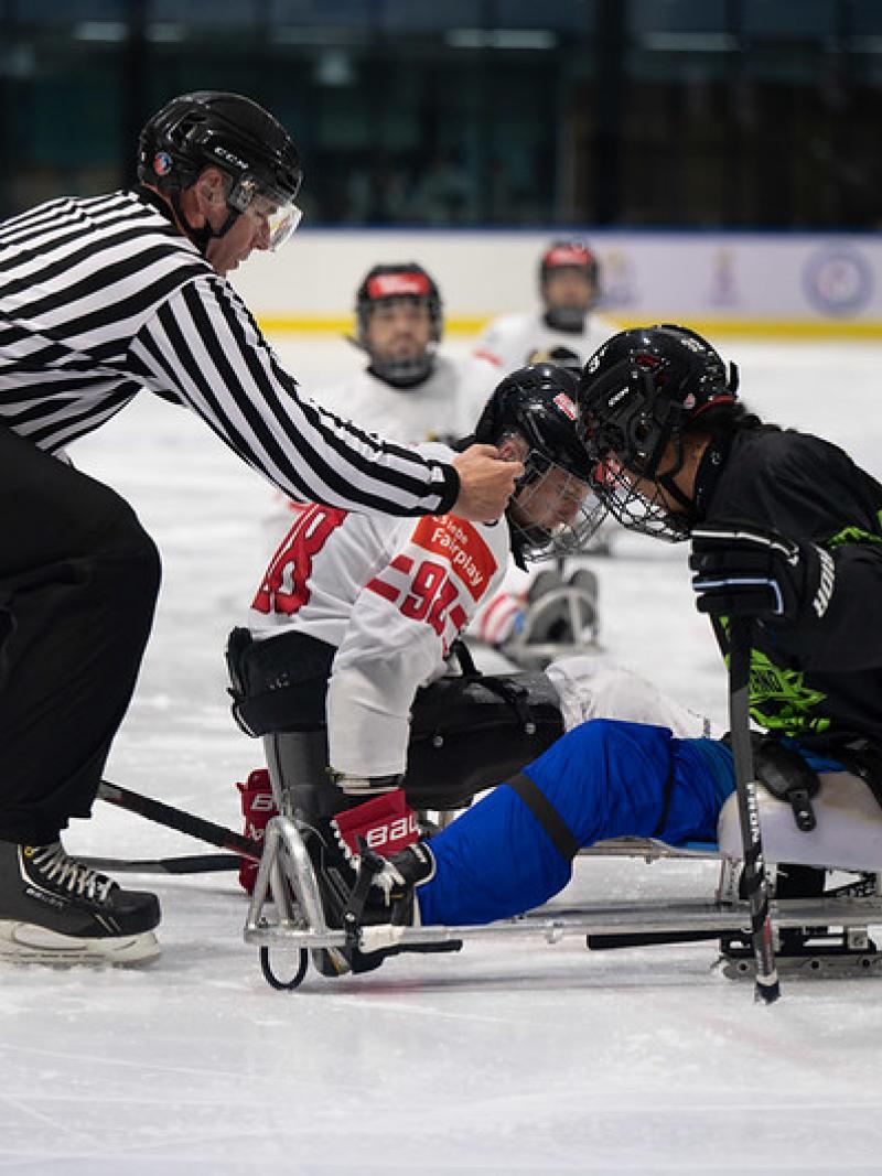 A Para ice hockey official dropping the puck in front of two players