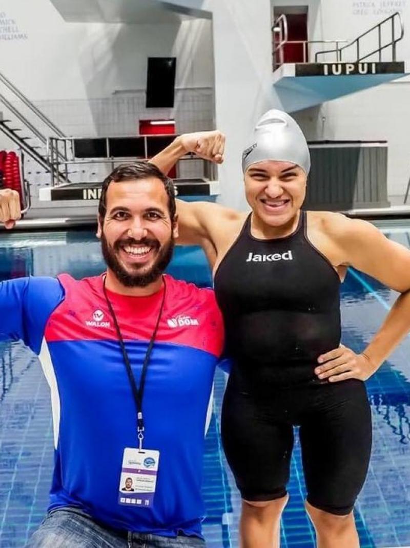 A short stature woman in swimming outfit with her arm around a knelled man with a swimming pool in the background