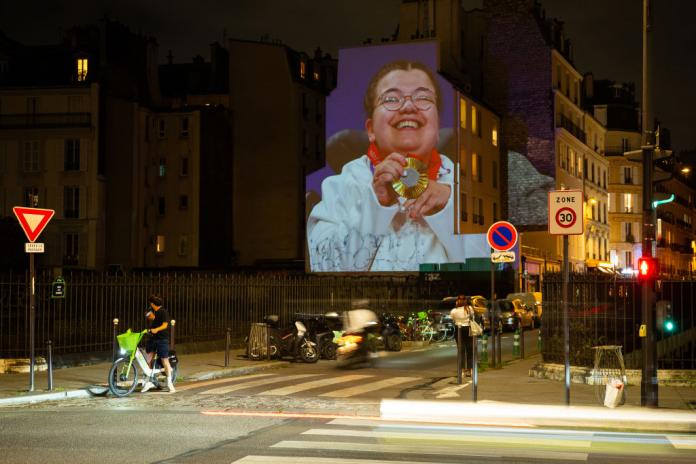 A photo of boccia athlete Aurelie Aubert smiling with her gold medal is projected on a building. There are pedestrians walking in front of the building