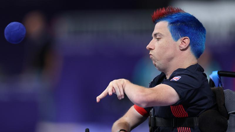 A man in a wheelchair with red and blue hair throws a blue boccia ball