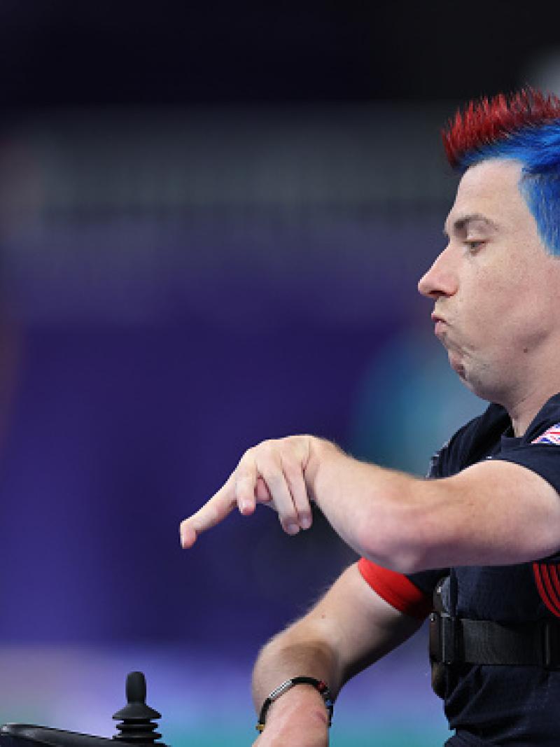 A man in a wheelchair with red and blue hair throws a blue boccia ball