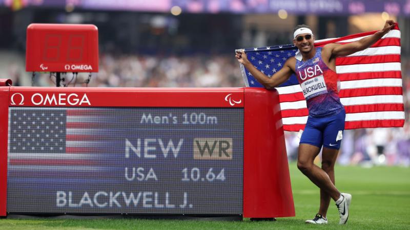 A man in a Team USA uniform holds up an American flag and stands next to an LED screen that shows his new world record time