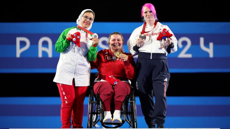 Three women pose on a podium, holding their medals
