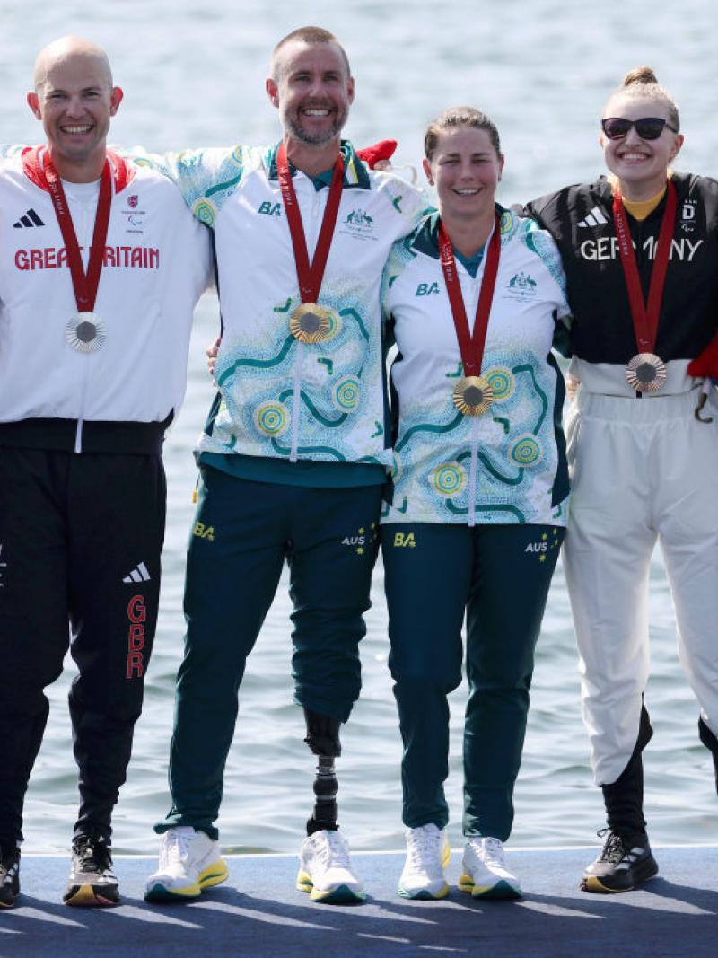 Six athletes stand on a podium next to water, two with silver medals, two with gold medals, and two with bronze medals