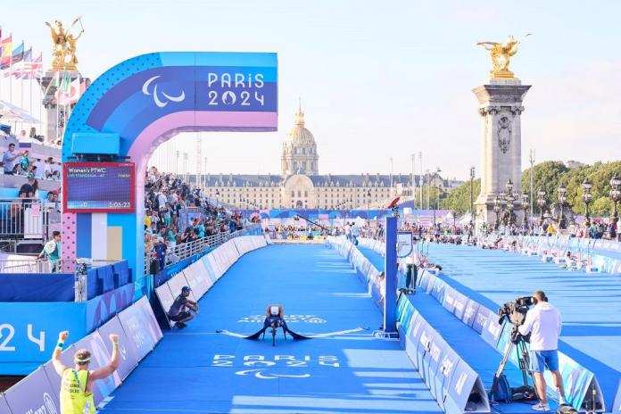 Bike crosses the para triathlon finish line with rider leaning forward showing a white helmet, surrounded by Paris 2024 branding
