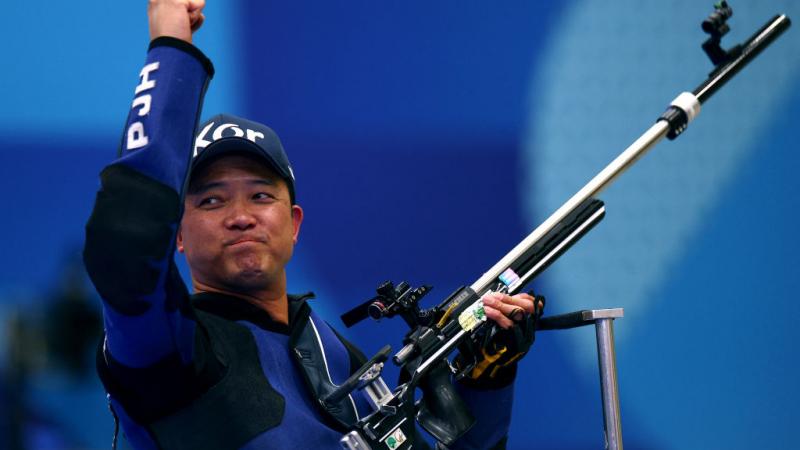A man in a dark blue jacket and baseball hat lifts one arm in the air in celebration and holds his shooting Para sport rifle in his other hand.