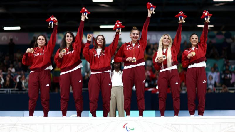 six women in red Turkiye uniforms hold up the Paris 2024 mascot and their gold medals with smiles on their faces