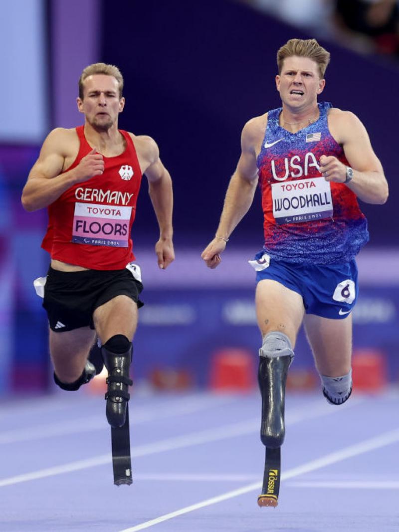 Three men with running blades race against each other, with the man in the middle, in a Team USA uniform, ahead of the other two