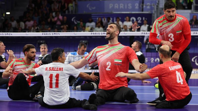 Five players wearing Team Iran uniforms yell in celebration during a sitting volleyball match