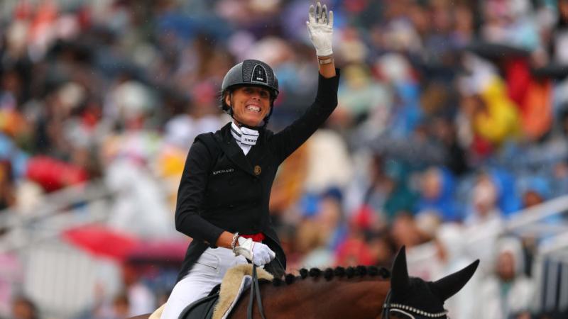 A female equestrian rider in a black jacket and helmet celebrates winning while sat on top of her horse