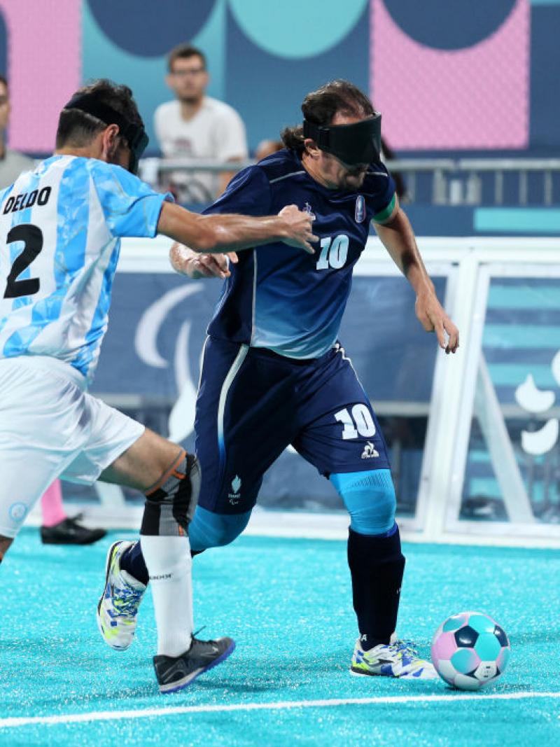 Two blind football players in Argentina uniforms and black eyeshades chase a French player, who has the ball