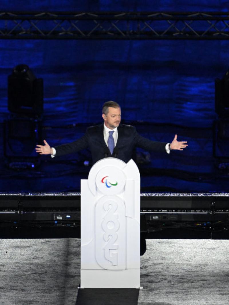A man on a stage raises his arms and stands behind a podium that is decorated with the year 2024 and the Paralympic agitos