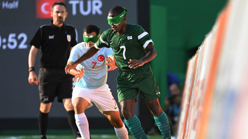 A man in green with a green blind fold tackles a man in blue taking the football away