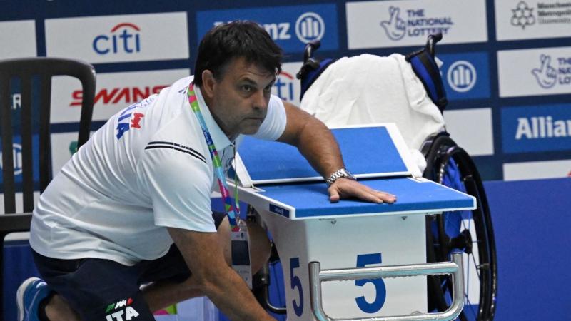 A male coach sits beside the pool and places his hand on a male swimmer's hand.