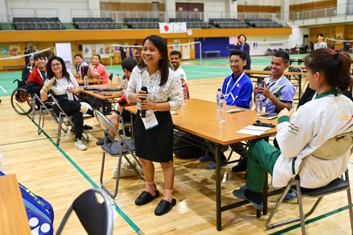 A woman is speaking in front of a group of athletes at a gymnasium