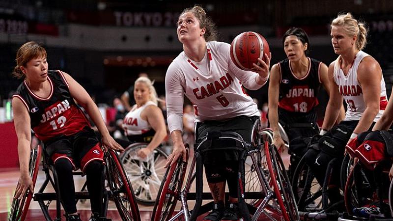 A female wheelchair basketball player carries the ball, while other players surround her